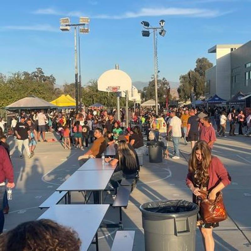 Crowded outdoor campus event with people socializing, long tables, tents, and a basketball hoop in the background