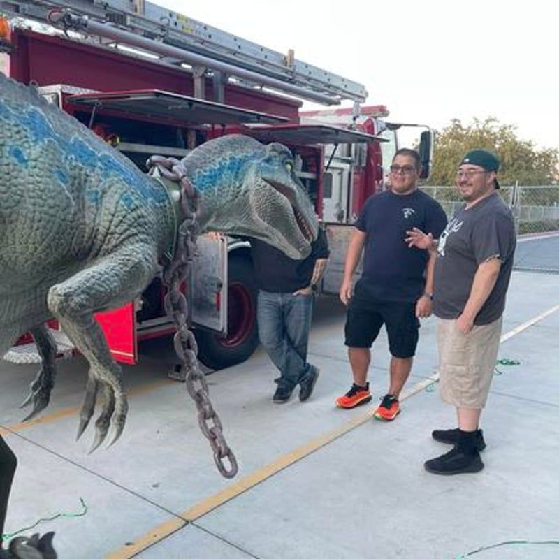 Two men standing beside a large blue dinosaur model near a red truck on a concrete lot