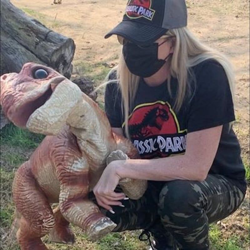Person in a Jurassic Park shirt and cap kneels beside a baby dinosaur prop outdoors.