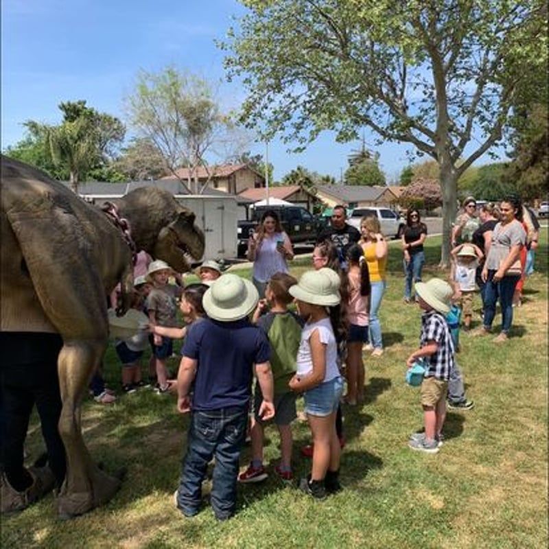 Children gather around a dinosaur exhibit outdoors while a handler speaks nearby.
