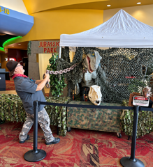 Man posing beside a dinosaur-themed display under a white canopy in a mall-like setting