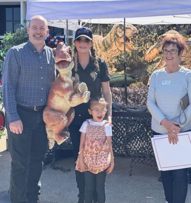 Four people posing with a standing dinosaur figure at an outdoor fair booth under a white tent.