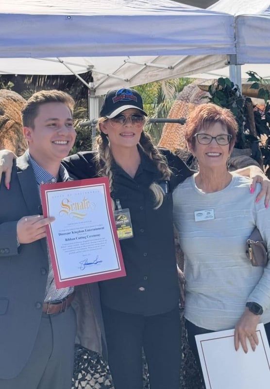 Three people smiling outdoors under a white canopy, holding certificates at a community event.