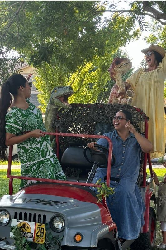 People posing on a red car in a park, with two dogs and green trees overhead
