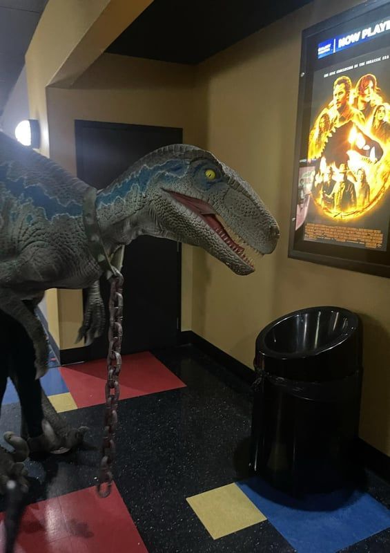 Blue dinosaur statue in a movie theater lobby beside a trash can and glowing movie poster.