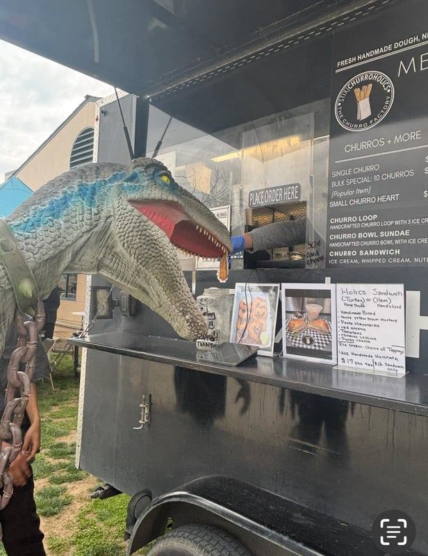 Large dinosaur sculpture beside a black food truck window with a menu and counter
