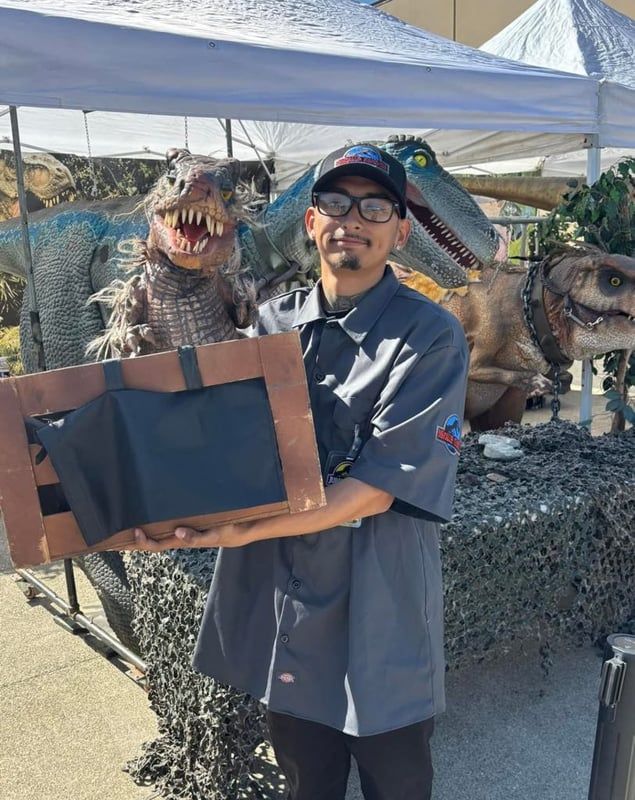 Man holding a framed sign beside dinosaur statues under a white canopy outdoors