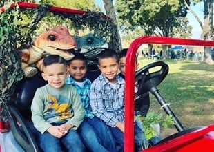 Three children sitting in a red open vehicle at a dinosaur-themed attraction