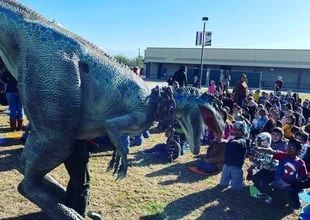 Life-size dinosaur puppet interacting with a crowd of children outdoors at a school event