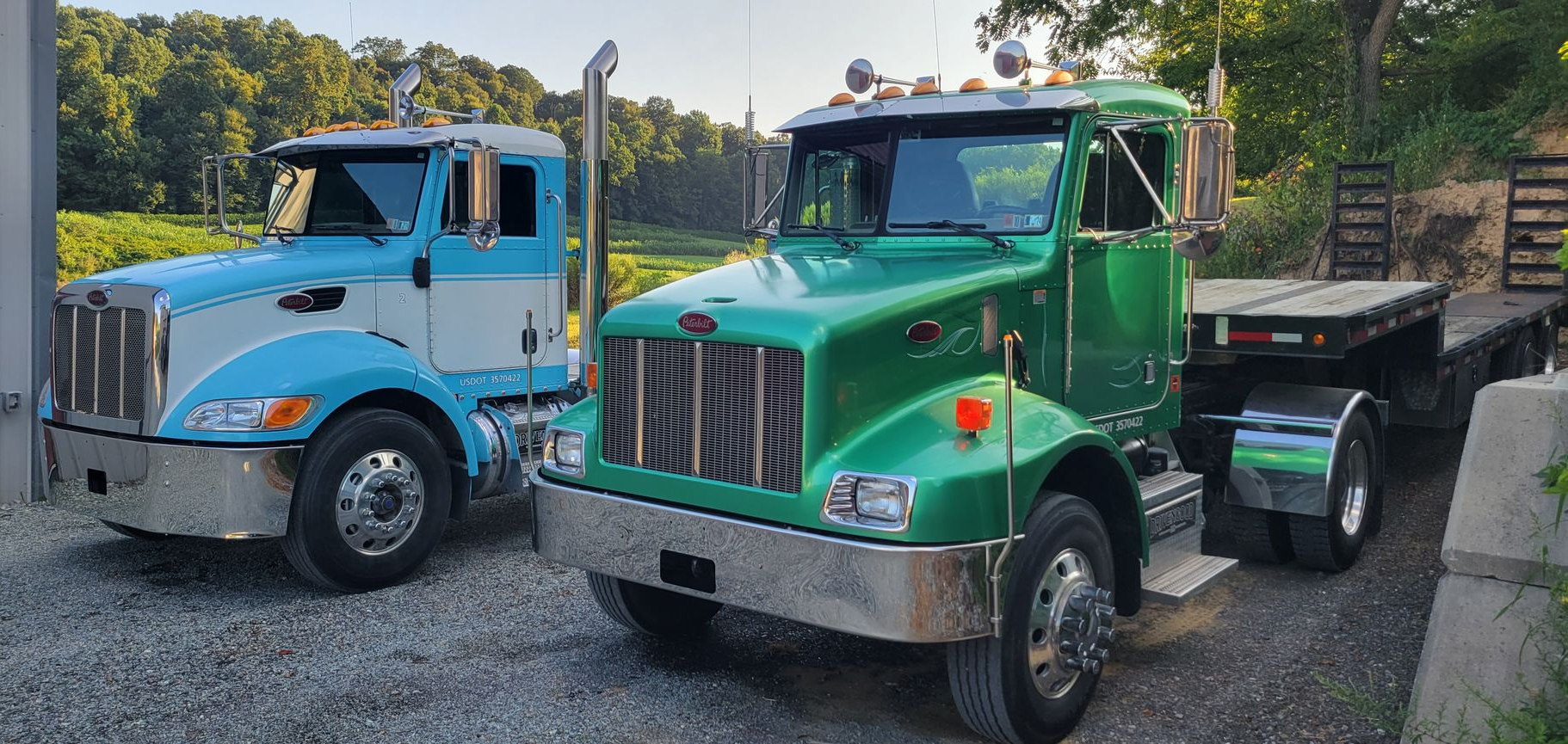 Two Peterbilt semi-trucks, one blue and white and one green, parked on a gravel lot near a wooded area.