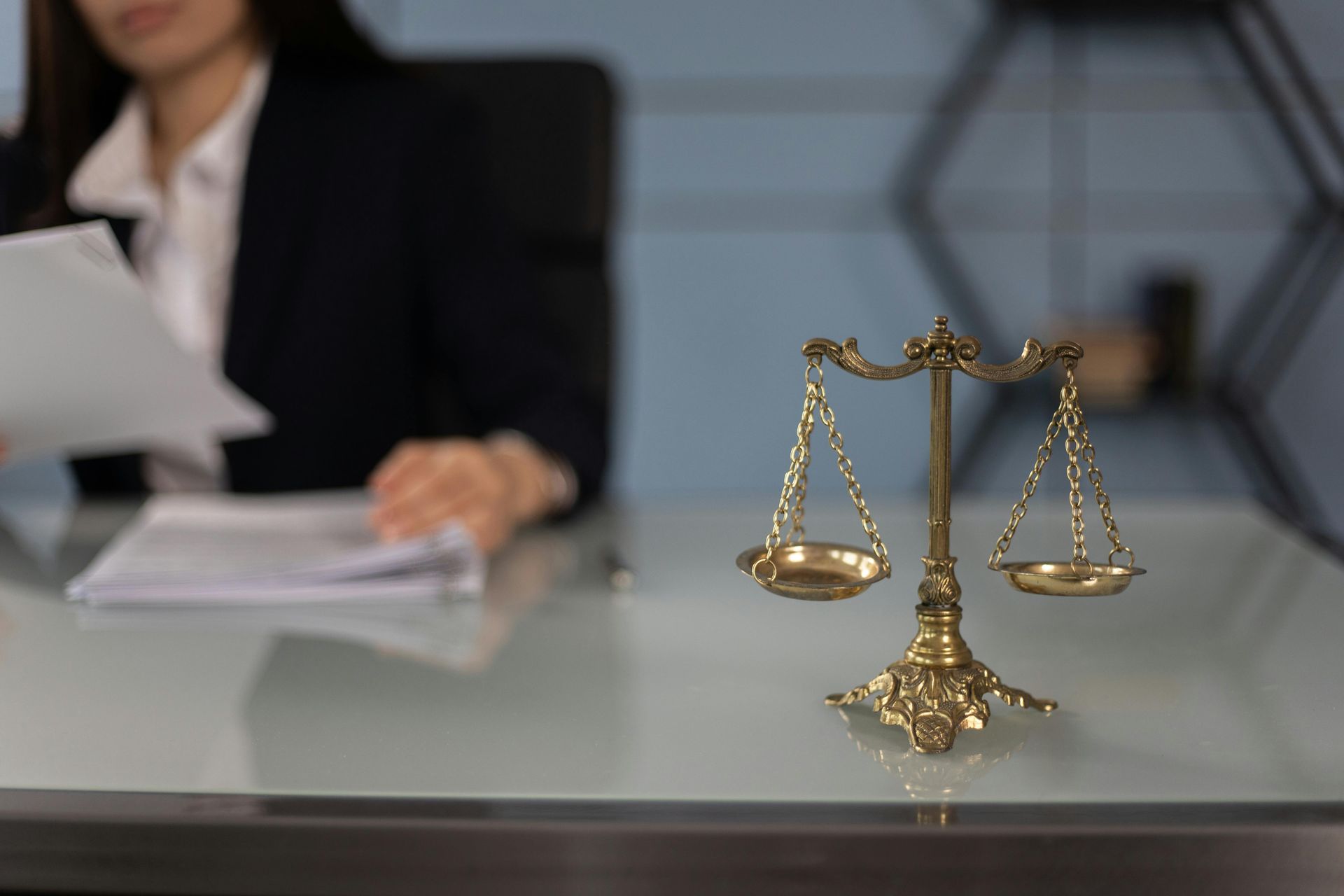 Gold scales of justice on a desk with a blurred person reviewing documents in the background
