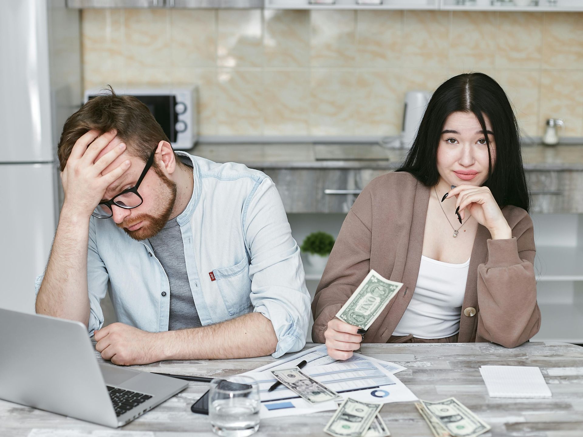 Frustrated person at laptop with bills and money, while another sits nearby at a kitchen table