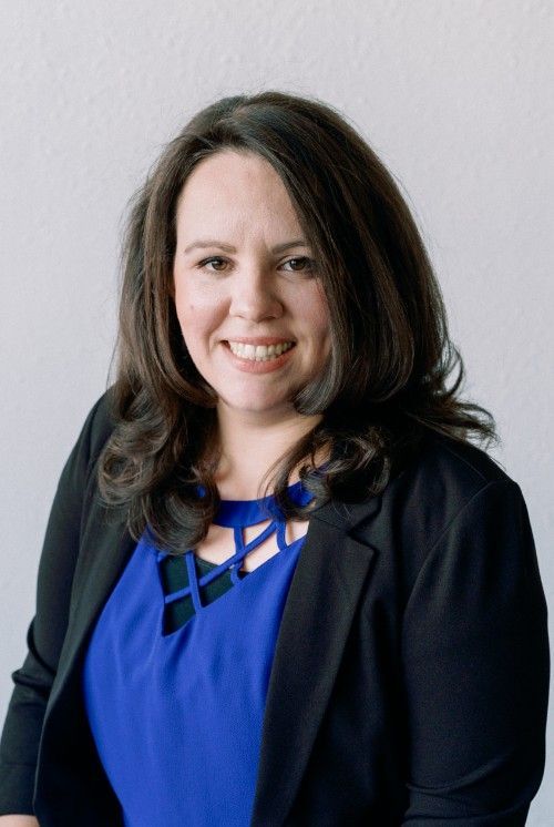 A woman in a blue dress and black jacket is smiling for the camera.
