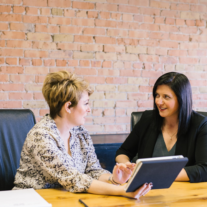 Two women are sitting at a table looking at a tablet.