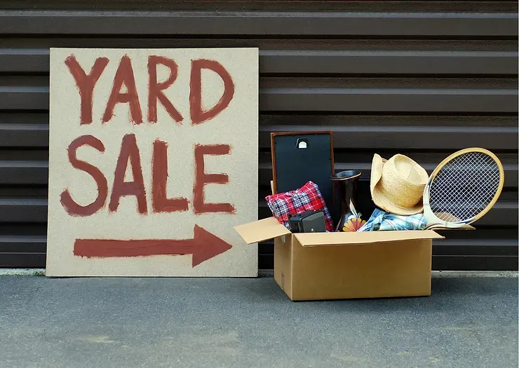 Yard sale sign with arrow, pointing to a cardboard box filled with items. Brown corrugated metal background.