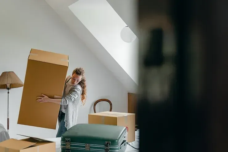 Woman carrying a large cardboard box in a room with other boxes, possibly moving.