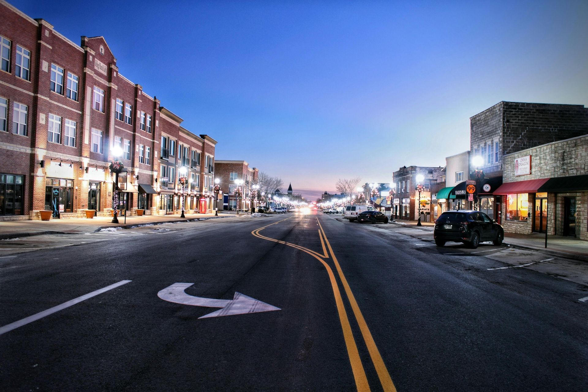 Street at dusk with buildings, streetlights, and a turn arrow painted on the road.