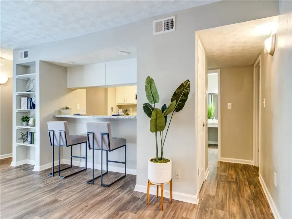 Modern open-concept apartment with kitchen bar stools, hallway, and potted plant on a light wood floor.