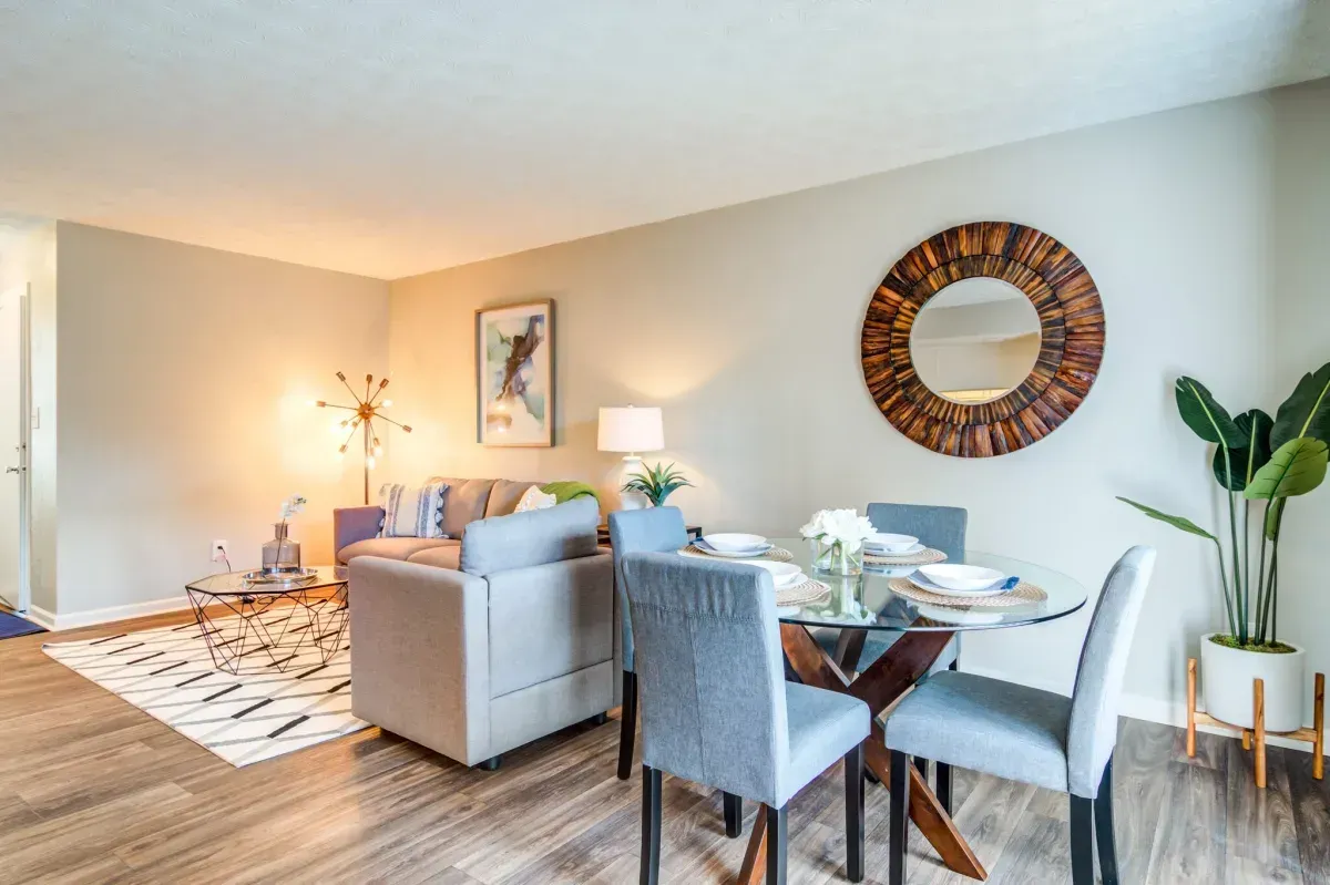 Bright living-dining room with gray furniture, wood floors, a round wall mirror, and a large plant.