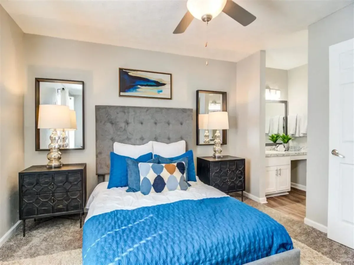 Bright bedroom with blue quilt, gray tufted headboard, mirrored nightstands, and ceiling fan