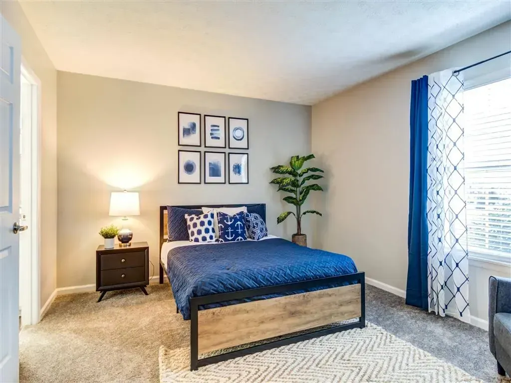 Bright bedroom with blue bedspread, bedside lamp, wall art, plant, and sliding glass door with curtains