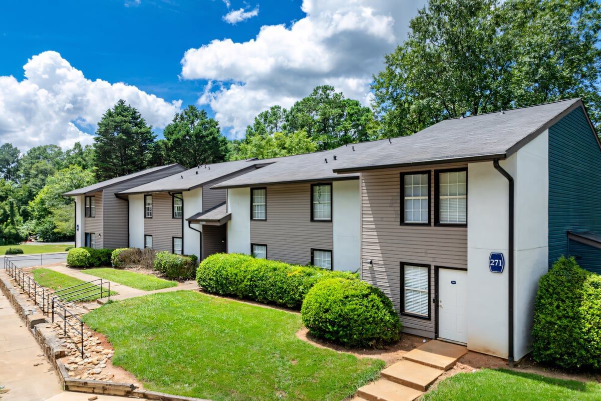 Row of beige apartment buildings with green lawns, shrubs, and a blue sky with clouds