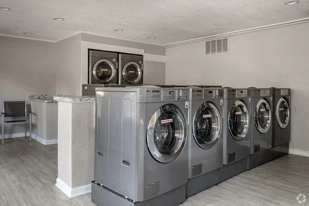 Laundry room with rows of gray front-loading washers and dryers, plus folding tables against a beige wall