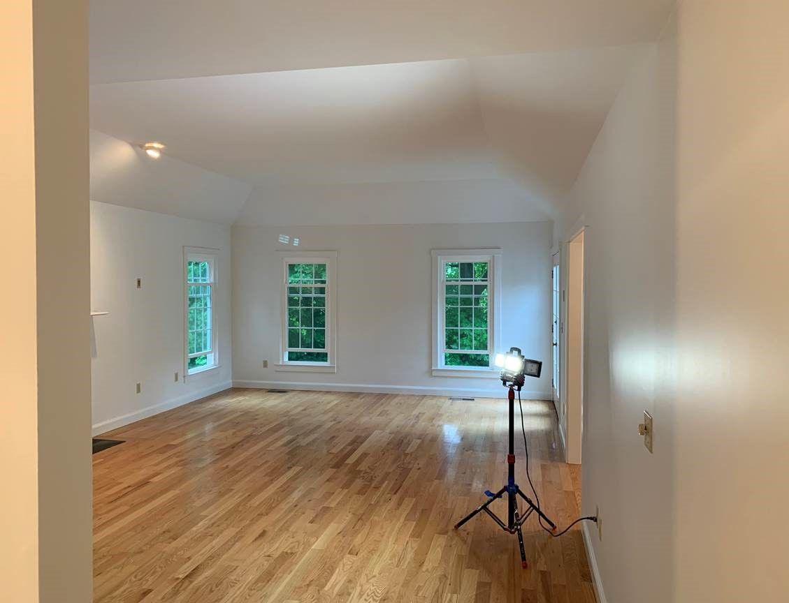Empty room with hardwood floors, white walls, three windows, and a light on a tripod stand.