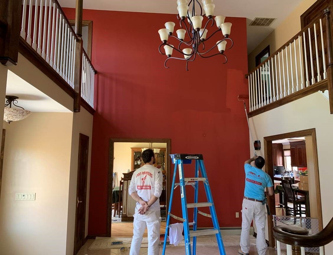 Two workers painting a red-walled foyer, with a blue ladder and chandelier overhead.