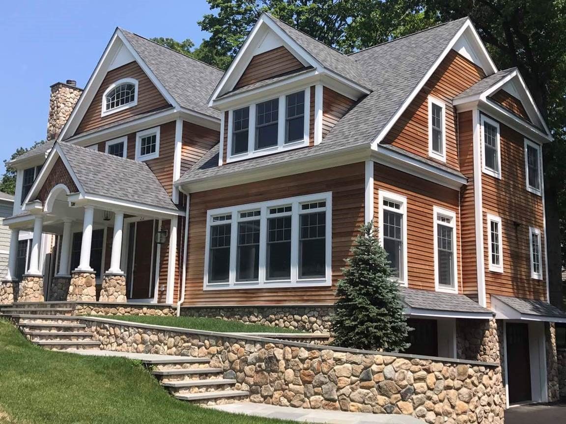 Large brick and wood house with stone steps, wraparound porch, and gabled roof on a grassy slope