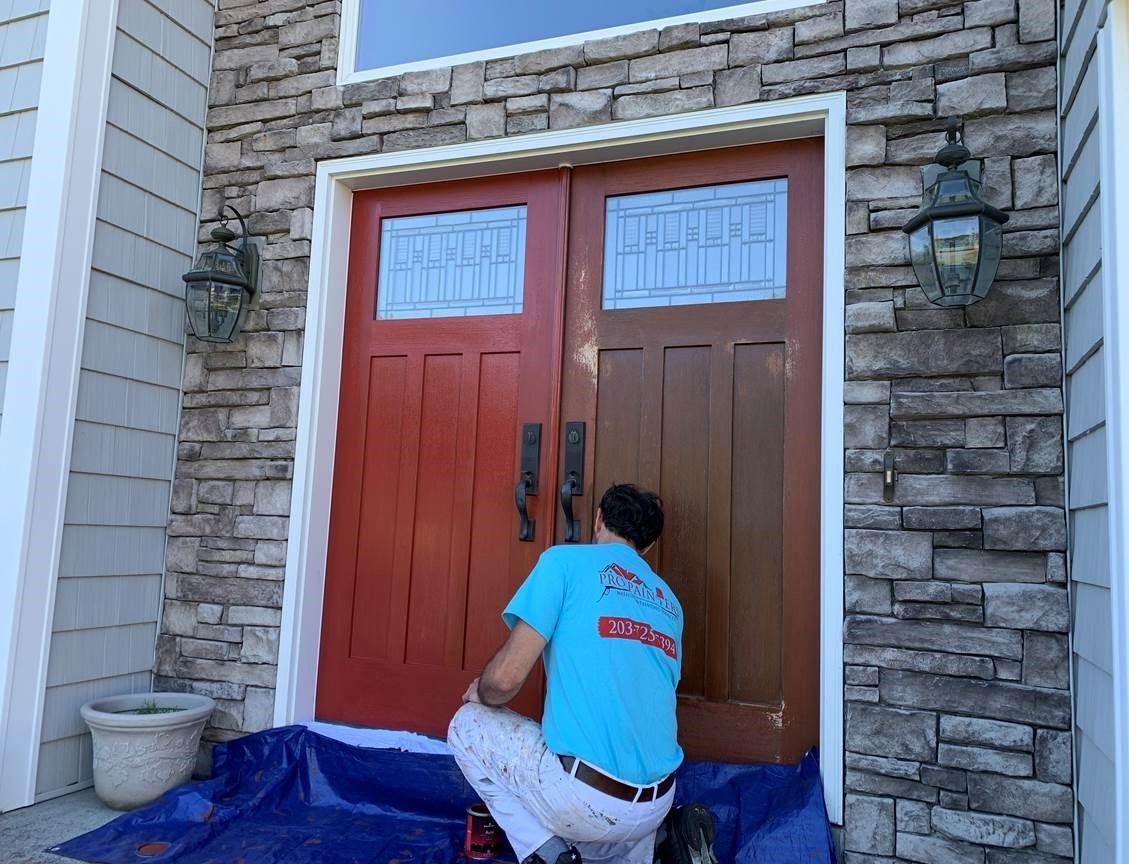 Person painting a brown front door next to a red one on a stone house exterior.