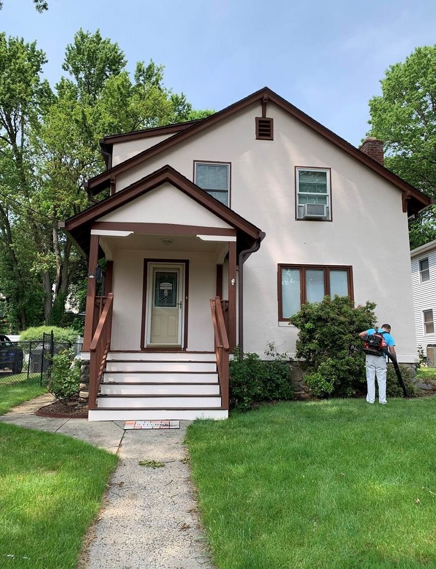 Two-story house with brown trim, front porch, green lawn, and a person standing by the right side.
