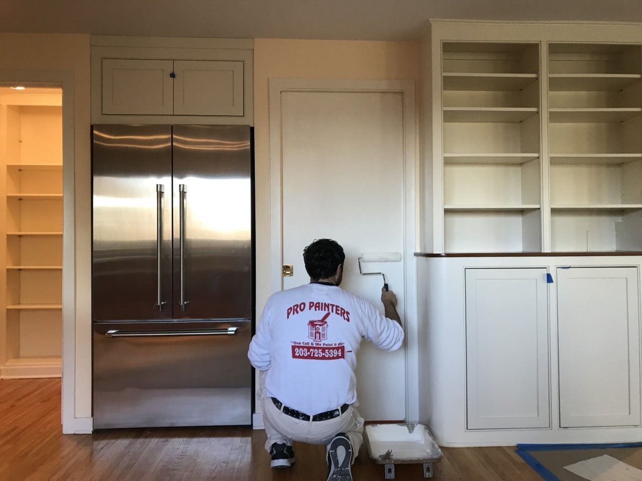 Person kneeling in a kitchen, painting a wall beside stainless steel refrigerator and white cabinets