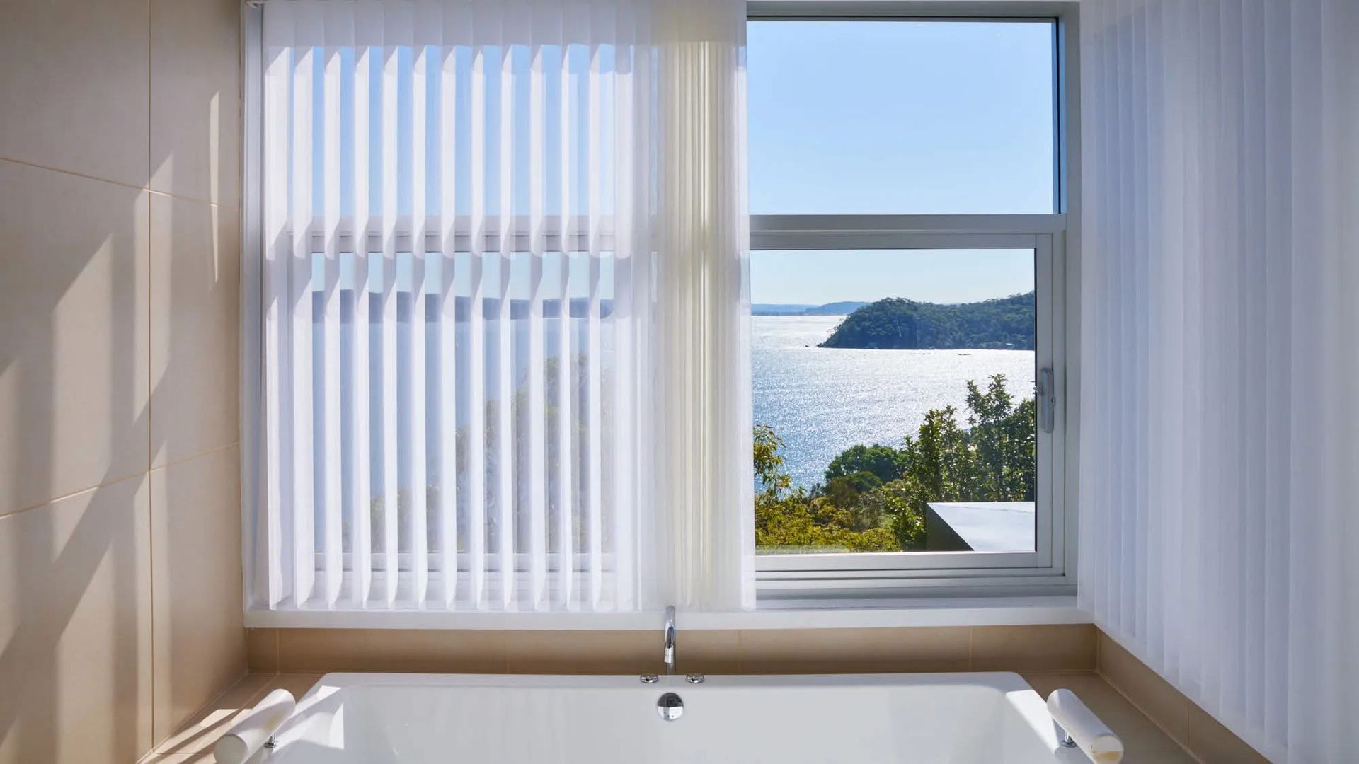 A bathroom with a bathtub and a window with a view of the ocean. — Phoenix Blinds In Albion Park Rail, NSW