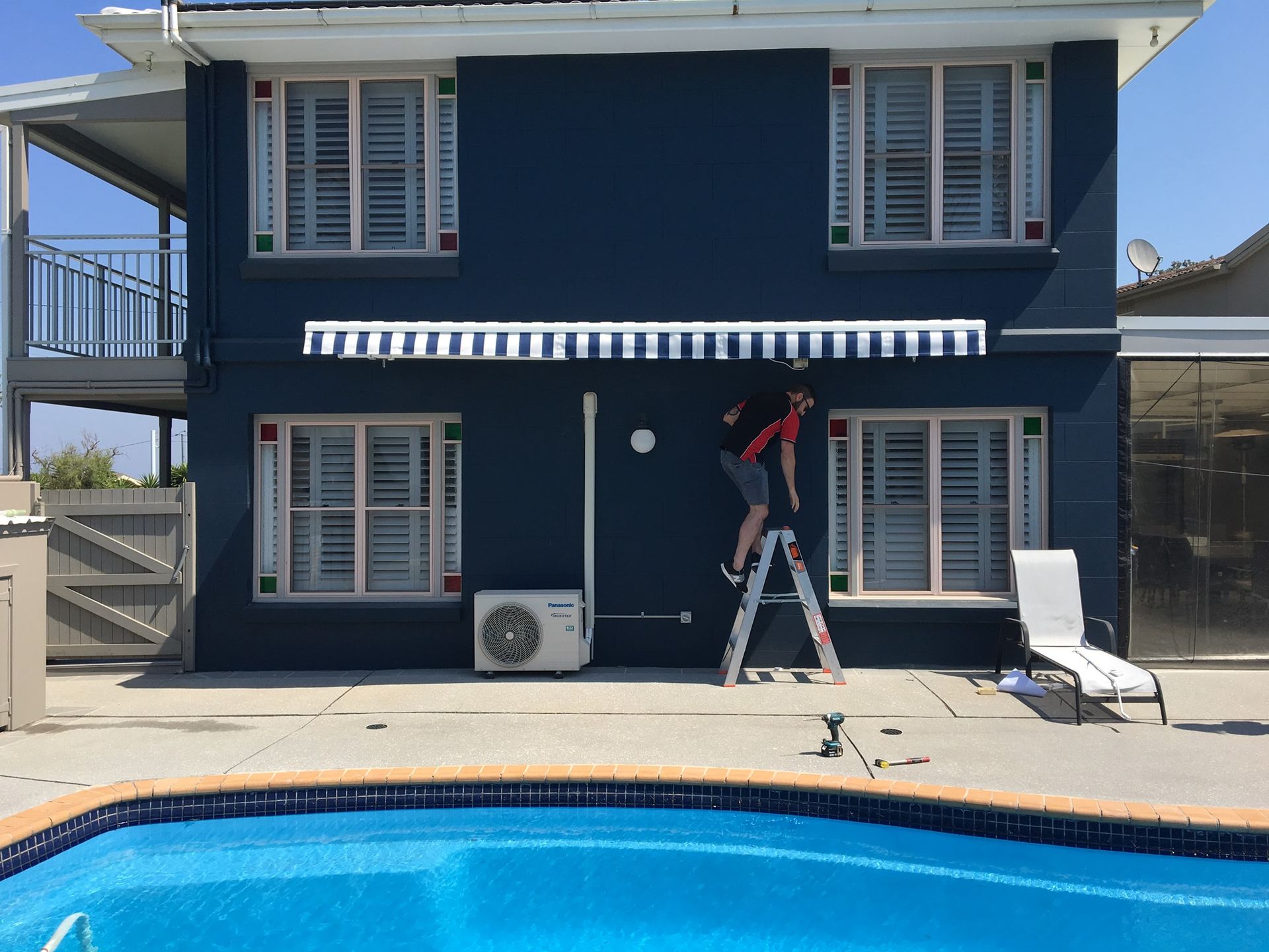 A Man Is Standing on A Ladder in Front of A Blue House Next to A Pool — Phoenix Blinds In Albion Park Rail, NSW