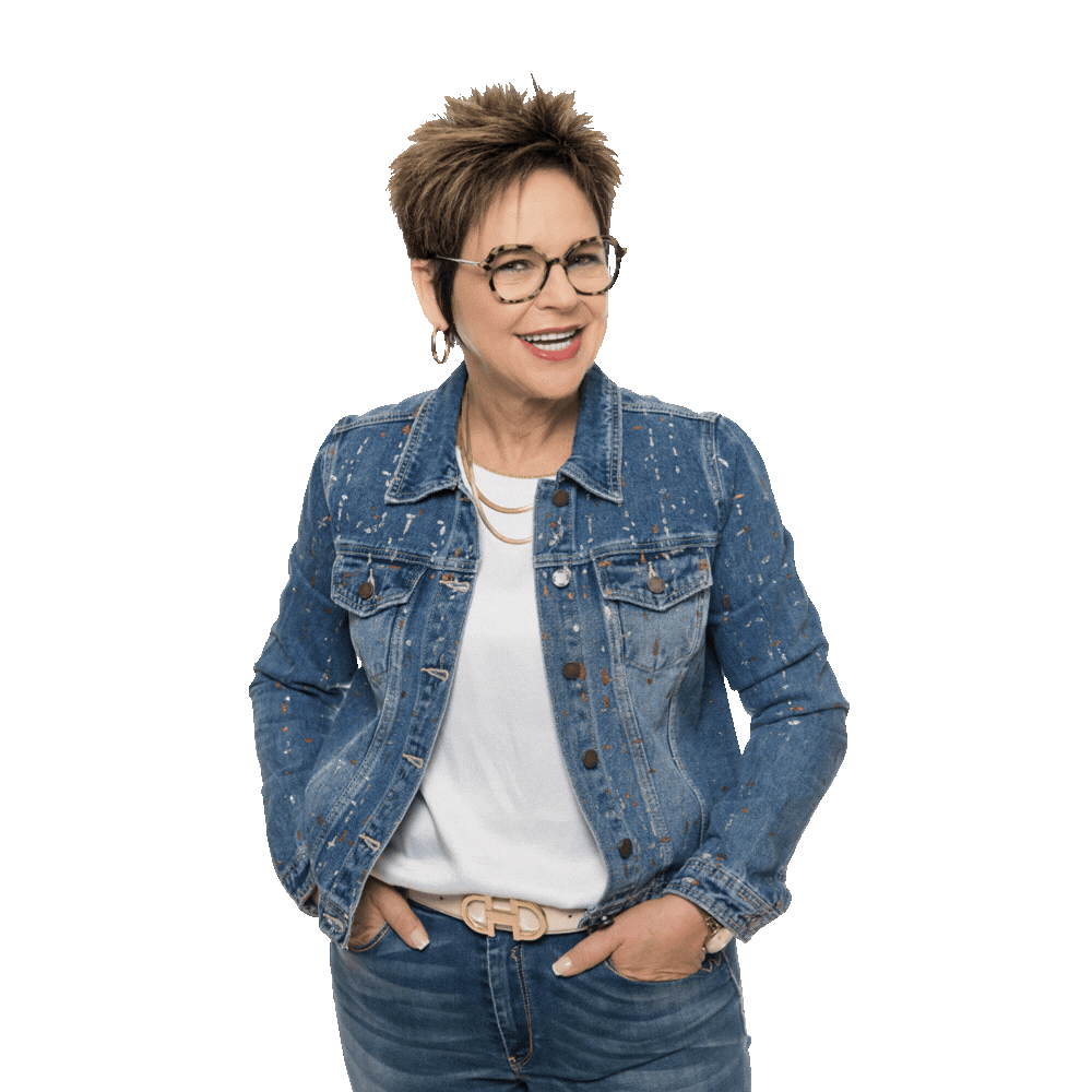 A smiling person with short brown hair wears glasses, a white t-shirt, and a blue denim jacket with hands in their pockets.