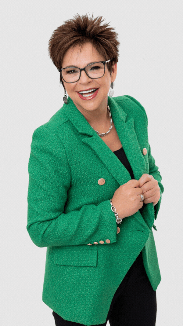 A person with short, dark hair and glasses smiles, wearing a green textured blazer over a black top against a gray background.