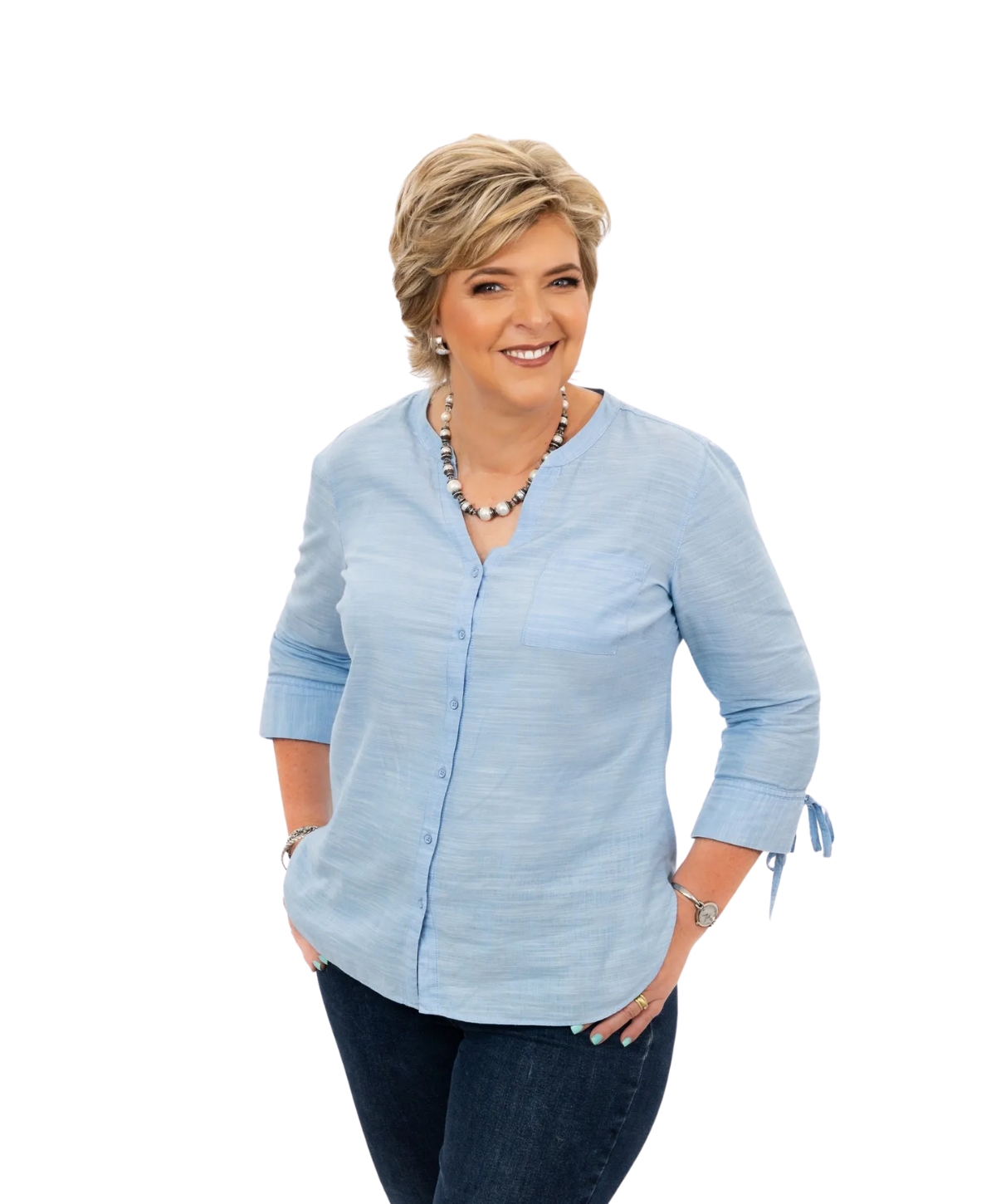 Smiling person wearing a light blue, button-down blouse and dark jeans, standing with hands in pockets against white.