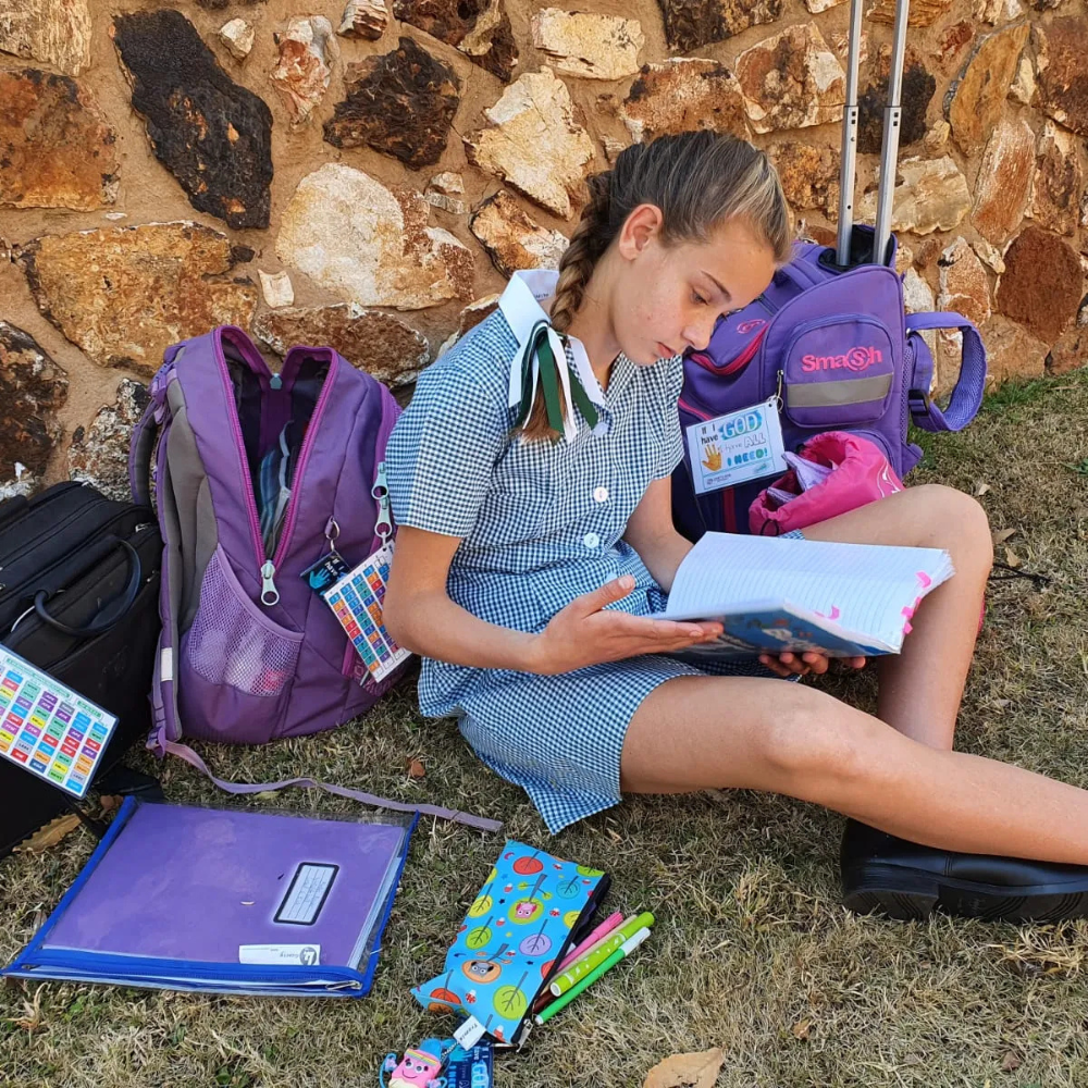 A student in a blue checkered uniform sits on grass reading a book, surrounded by school bags and supplies.