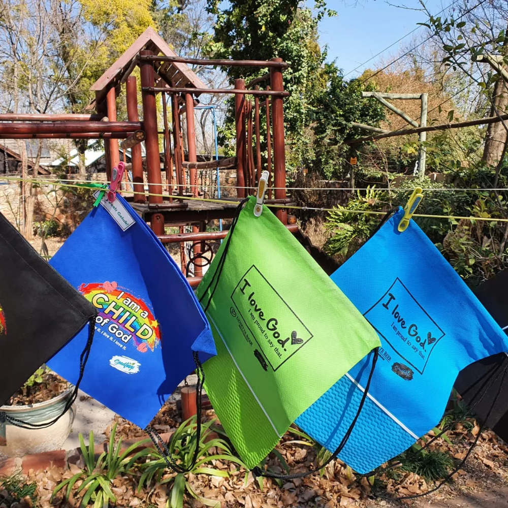 Bright blue and green drawstring bags with religious text hanging on a clothesline outdoors in front of a wooden play set.