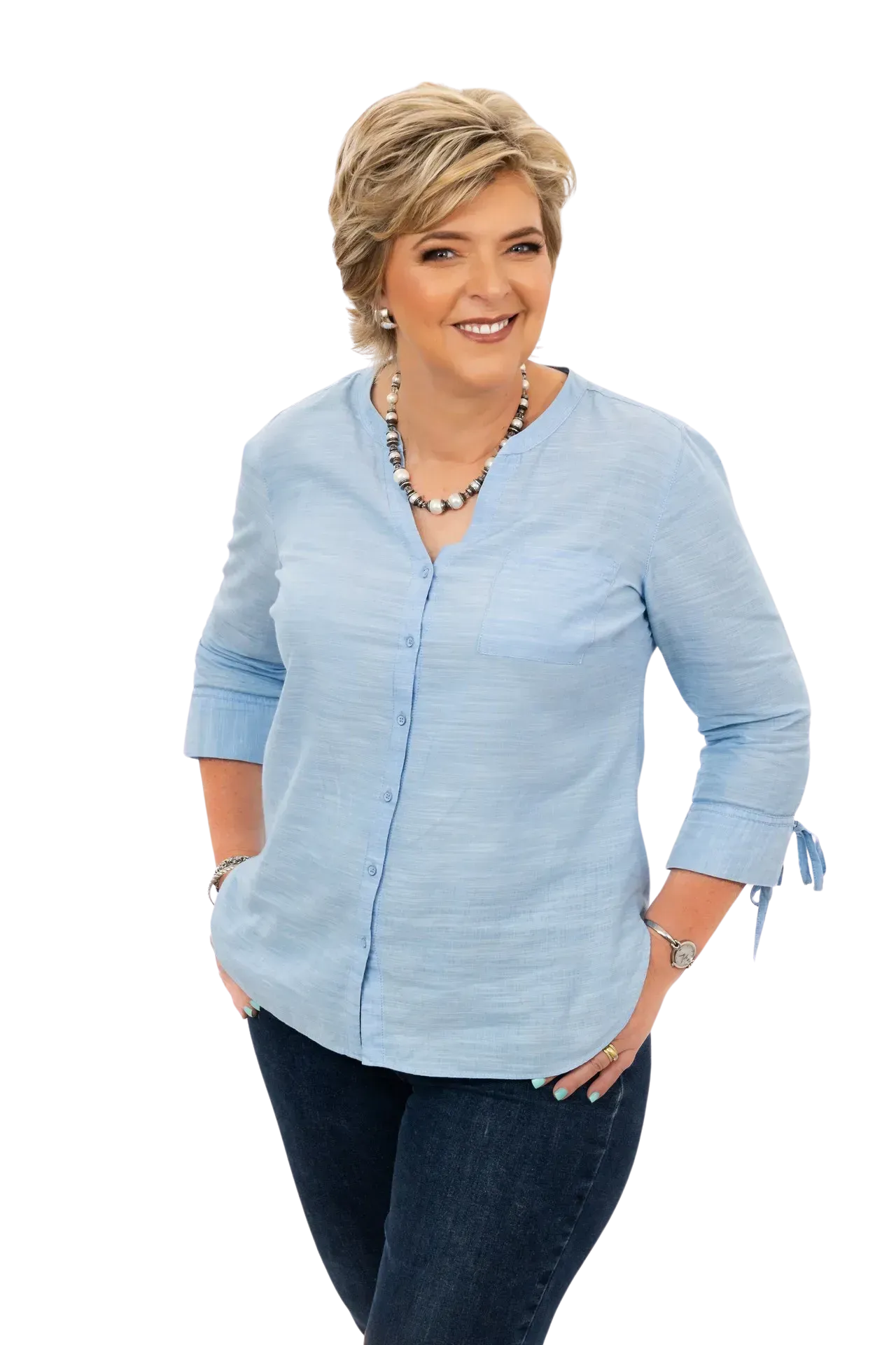 Smiling person wearing a light blue, button-down blouse and dark jeans, standing with hands in pockets against white.