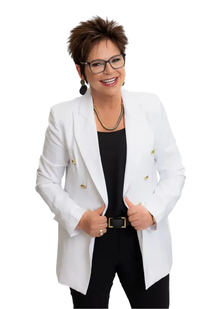 A person with short dark hair and glasses wears a white blazer over a black top, smiling while posing against a white background.