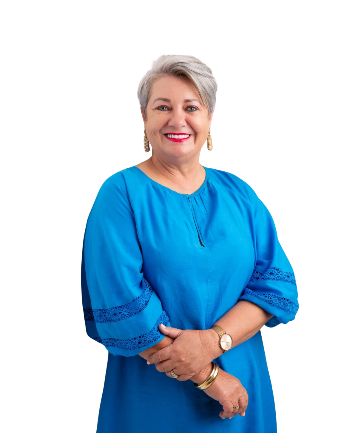 A smiling person with short, light hair wearing a bright blue long-sleeved dress and gold jewelry, arms crossed.