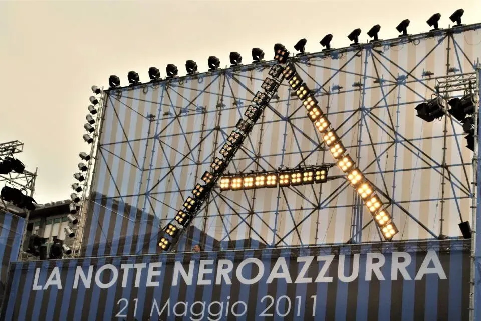 Ponteggio illuminato a forma di A sulla facciata di un edificio con le scritte “LA NOTTE NEROAZZURRA” e “21 Maggio 2011”.