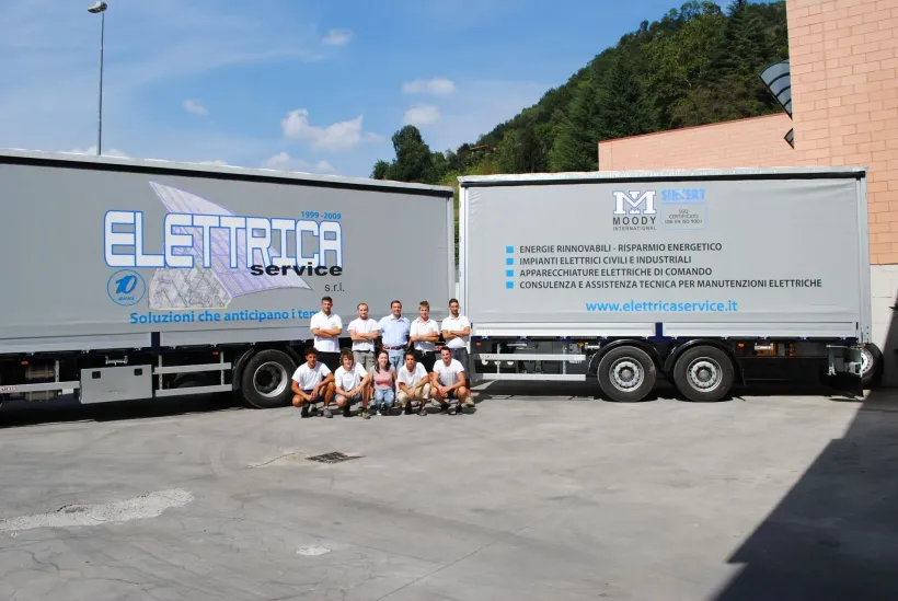 Operai in posa davanti a due camion bianchi della ELETRIC in un parcheggio soleggiato.