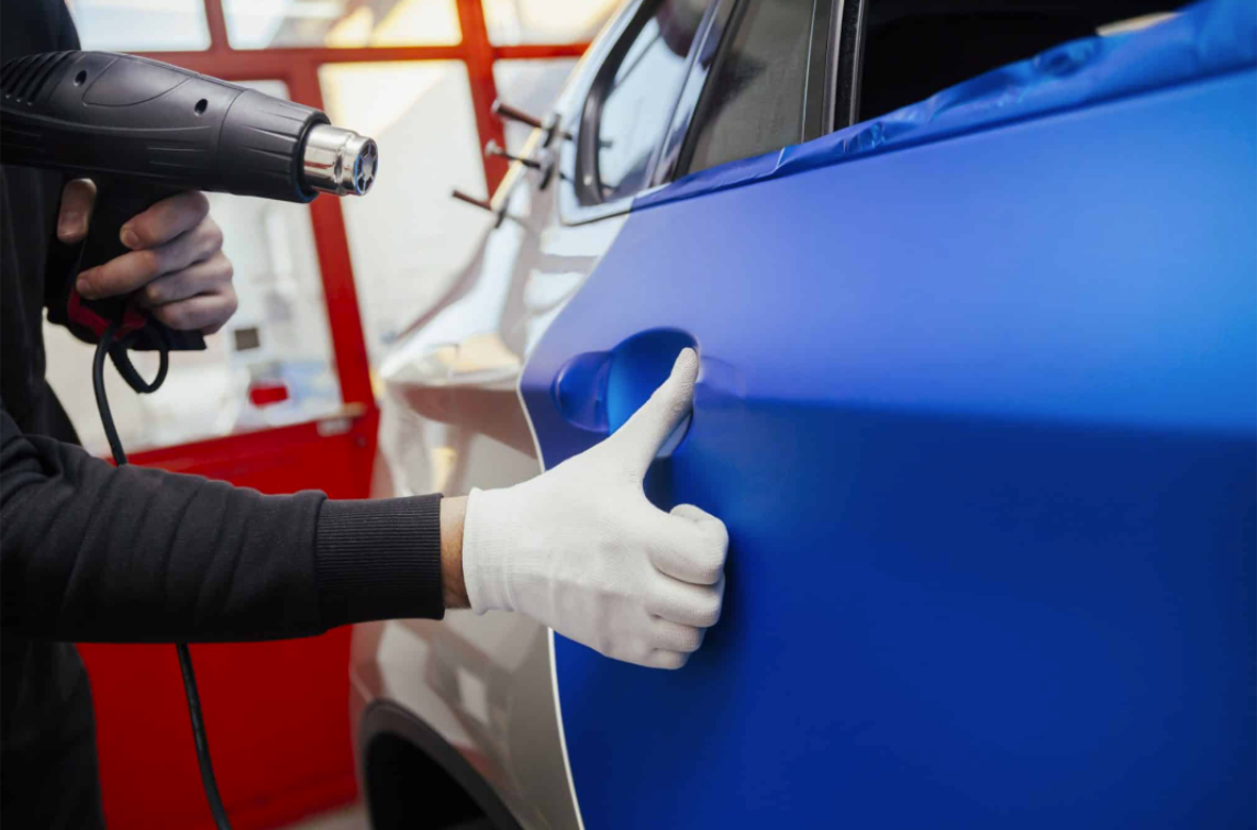 Person using a heat gun to apply blue vinyl wrap to a car door, wearing gloves.