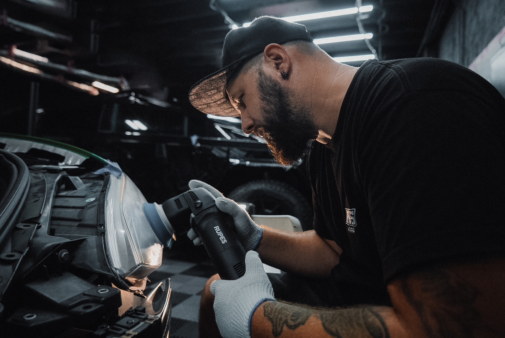 Man polishes car headlight with a buffing tool. He's wearing a cap, gloves, and a black shirt in a garage.