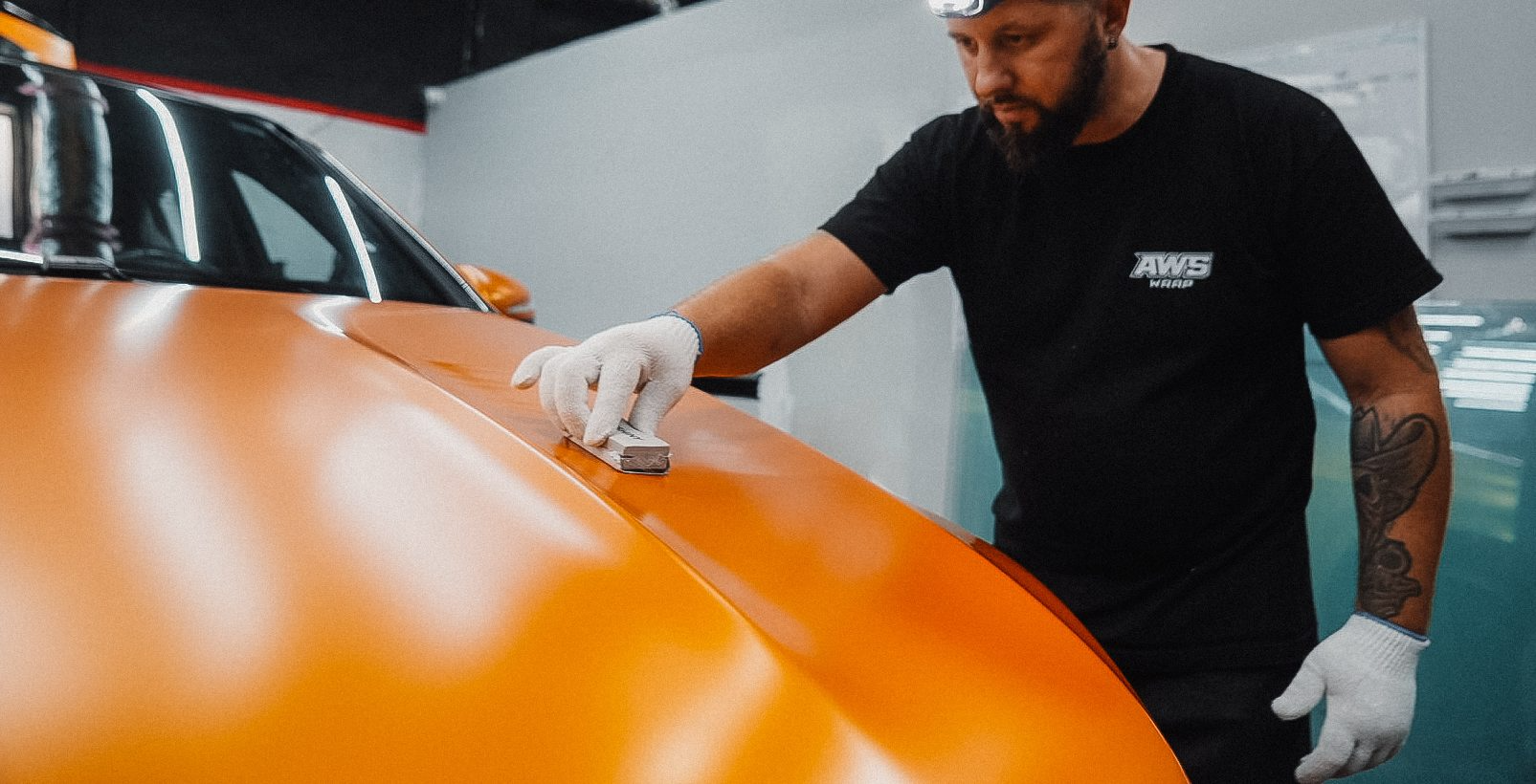 Man applying orange vinyl wrap to a car hood, wearing gloves and a headlamp in a shop.