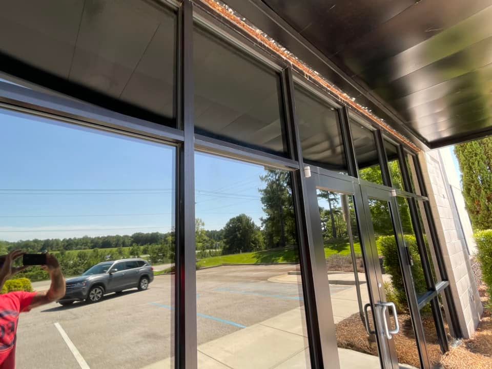 Glass-fronted building exterior reflects sky, car, and person taking a photo. Sunny day; black window frames.