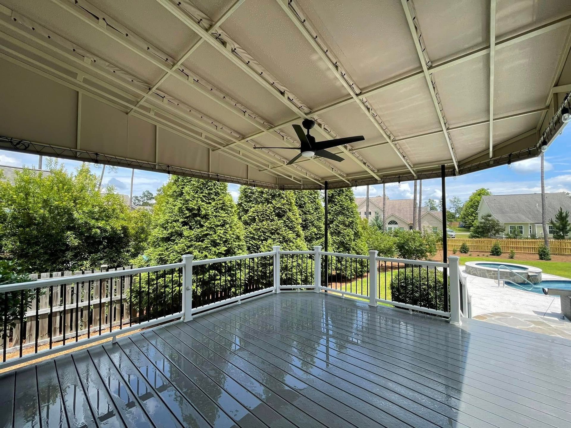 Covered outdoor deck with grey flooring, black railing, and a ceiling fan; trees and a pool in the background.